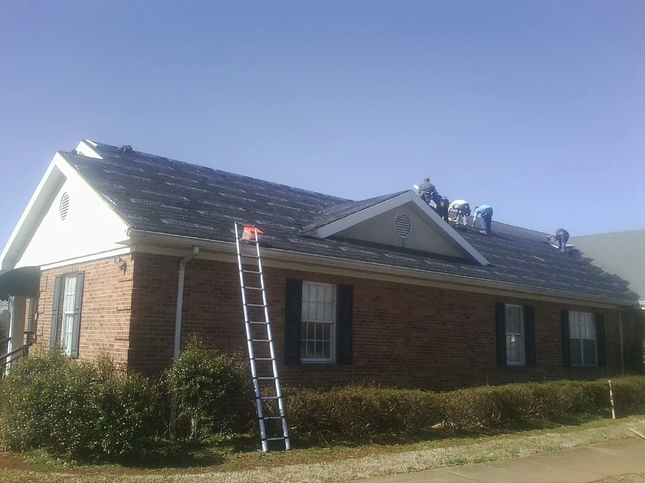 Crew tearing shingles off a brick ranch roof with a ladder set — Emergency Roof Repair work by our crew in East Flat Rock