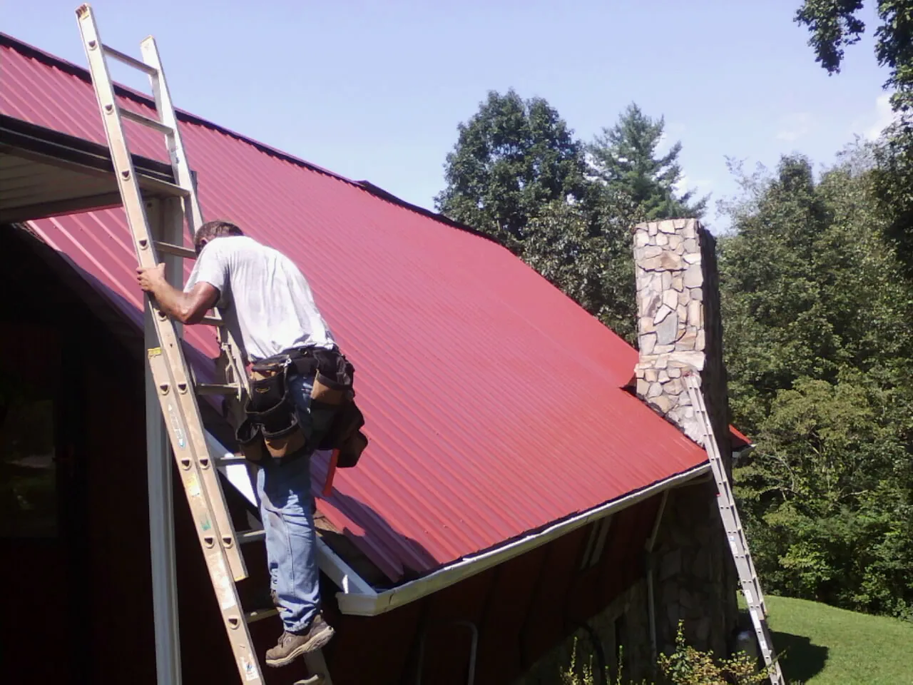 Roofer on a ladder beside a red metal roof and stone chimney — Soffit & Fascia Repair by Roofing Brothers in East Flat Rock